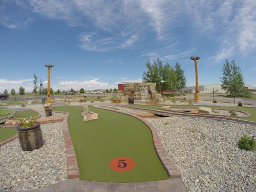 A mini-golf course featuring green turf, rocky paths, and a stone structure under a blue sky with scattered clouds.