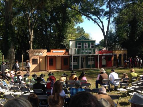 A crowd watches a Western-themed performance in front of colorful buildings in a park setting.