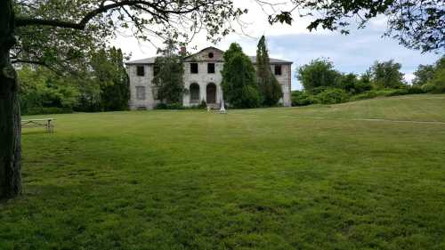 An old, abandoned building surrounded by greenery and a large lawn under a cloudy sky.