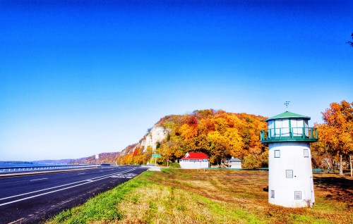 A scenic view of a highway with a lighthouse and colorful autumn trees on a hillside under a clear blue sky.