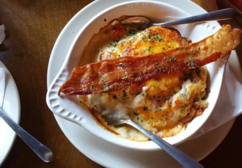 A creamy dish topped with crispy bacon and herbs, served in a white bowl with two spoons on a wooden table.