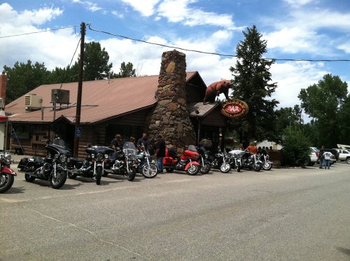A rustic bar with a stone chimney, surrounded by parked motorcycles and a bear sign, under a blue sky.
