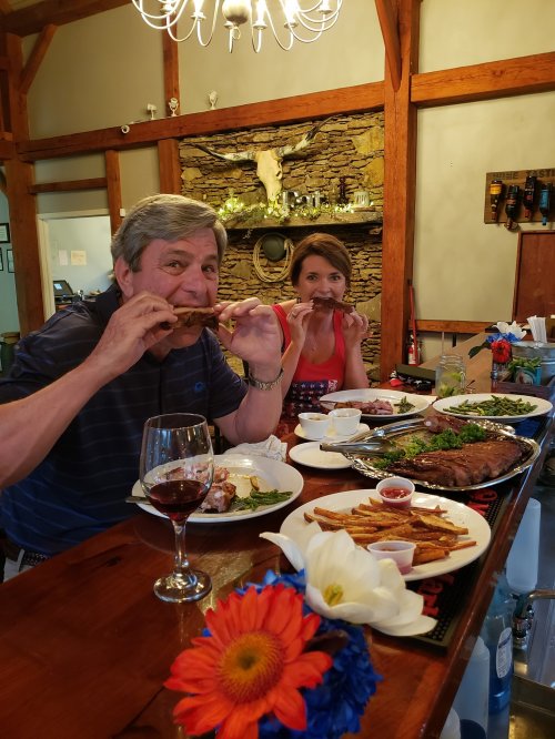 A man and woman enjoy a meal at a rustic table, featuring steak, fries, and wine, with flowers in the foreground.