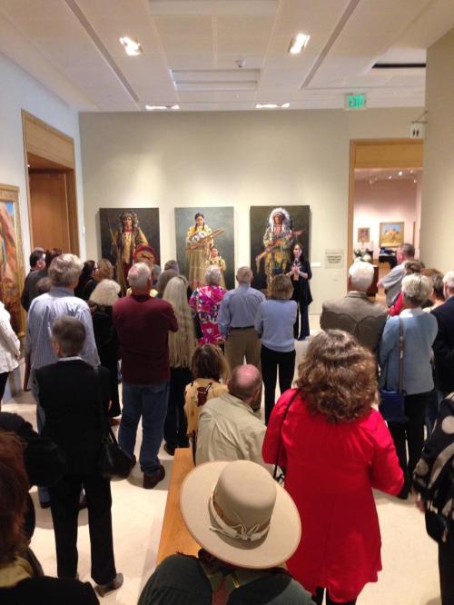 A crowd of people gathers in an art gallery, listening to a speaker in front of large paintings.