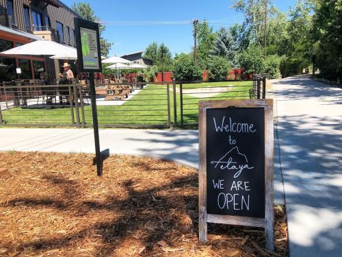 A sign reading "Welcome to [Location], WE ARE OPEN" stands outside a restaurant with outdoor seating and greenery.