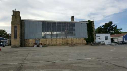 A large, industrial building with a glass front, surrounded by a paved area and trees in the background.