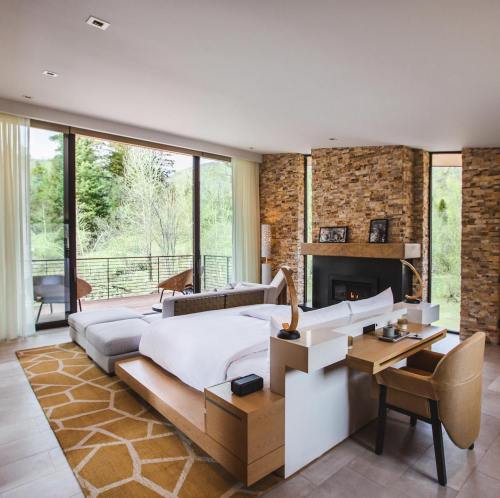 Modern bedroom with large windows, stone walls, a cozy bed, and a view of greenery outside. Stylish and serene atmosphere.