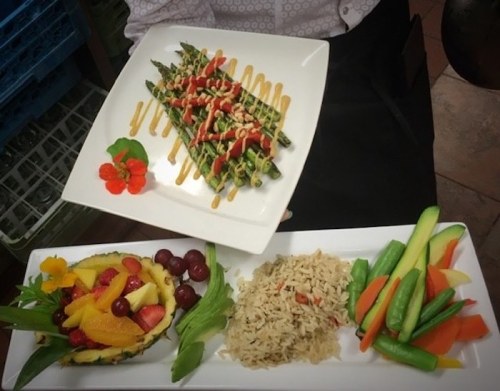 A plate of asparagus drizzled with sauce, garnished with flowers, alongside a fruit salad and rice with mixed vegetables.