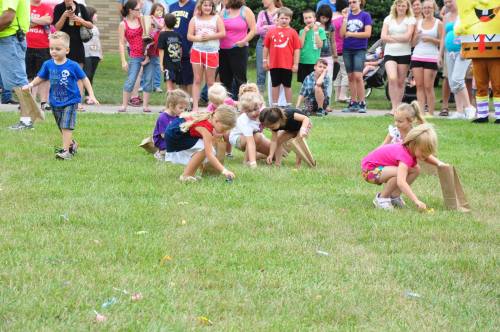 Children participate in a sack race on grass, while a crowd watches in the background.