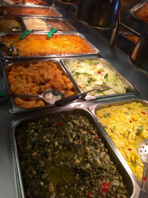 A buffet display featuring various dishes, including baked casserole, greens, mixed vegetables, and diced fruit.