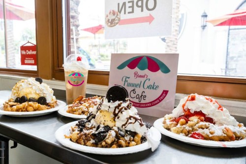 Four plates of funnel cakes topped with ice cream, whipped cream, and various toppings, with a sign for the café in the background.