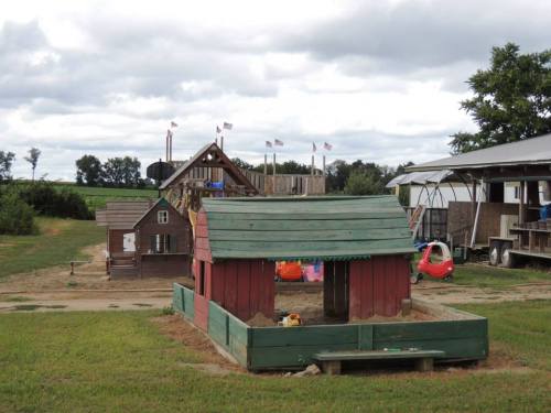 A colorful playhouse in a grassy area, with a larger play structure and trees in the background under a cloudy sky.
