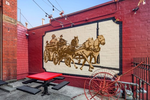Mural of a horse-drawn carriage on a red brick wall, with a red picnic table in front and vintage wheels nearby.