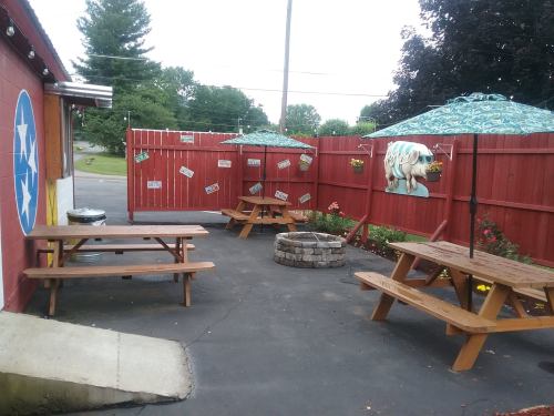Outdoor seating area with wooden picnic tables, umbrellas, and a fire pit, surrounded by a red fence and decorative elements.