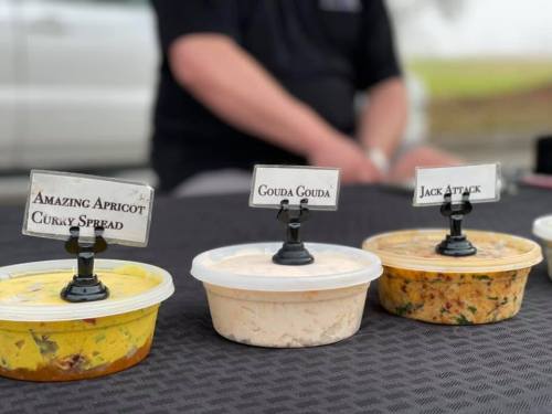 Three containers of flavored spreads labeled "Amazing Apricot Curly Spread," "Gouda Gouda," and "Jack Attack" on a table.