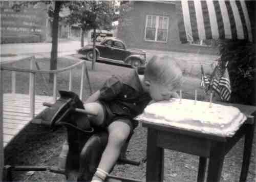 A young boy leans over a birthday cake with candles, sitting on a toy horse, with flags and a vintage car in the background.