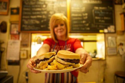 A woman in a red shirt holds a plate with two burgers and fries in a cozy diner setting.