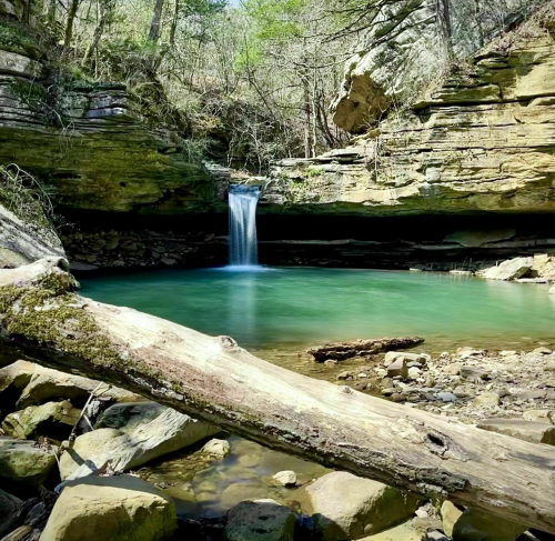 A serene waterfall cascades into a turquoise pool, surrounded by rocky cliffs and lush greenery.