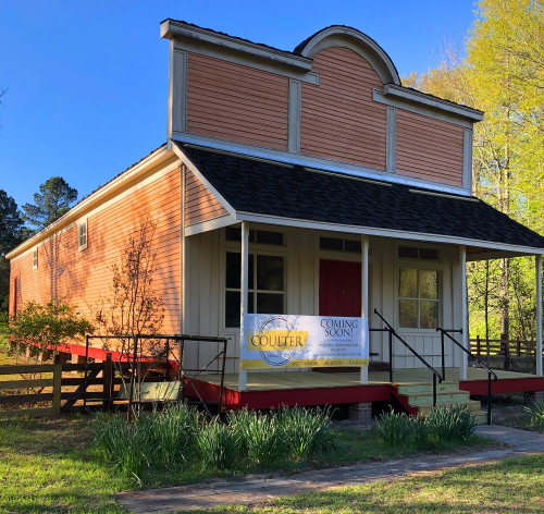A charming, two-story building with a porch, featuring a "Coming Soon" sign and surrounded by greenery.