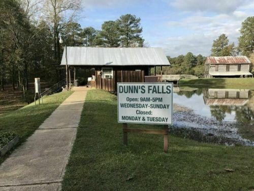 Sign for Dunn's Falls with hours of operation, surrounded by greenery and a pond, under a cloudy sky.