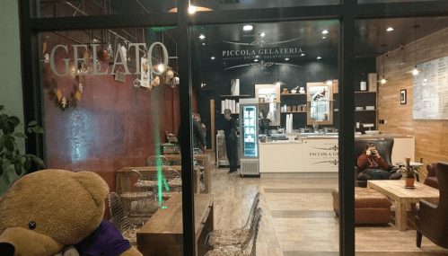 A cozy gelato shop with a large teddy bear outside, featuring a modern interior and a display of gelato flavors.