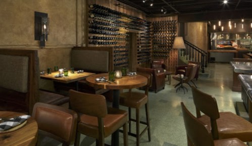 Cozy restaurant interior with wooden tables, leather chairs, and a wall of wine bottles in a warm, inviting atmosphere.