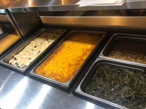 A buffet display with trays of various dishes, including baked casseroles and greens, on a stainless steel counter.