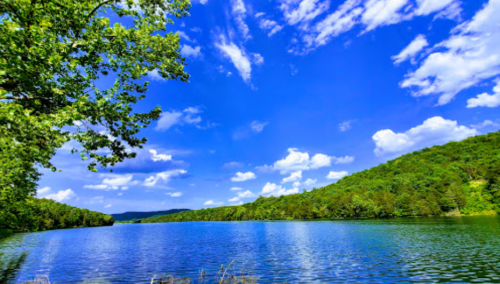A serene lake surrounded by lush greenery and rolling hills under a bright blue sky with fluffy white clouds.