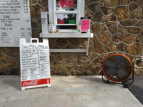 A menu board outside a small food stand with a stone wall, featuring drinks and snacks, next to a large fan.