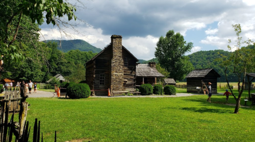 A rustic stone house surrounded by greenery, with mountains in the background and people exploring the area.
