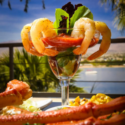 A shrimp cocktail in a glass, garnished with lettuce and tomato, with a scenic view of water and palm trees in the background.