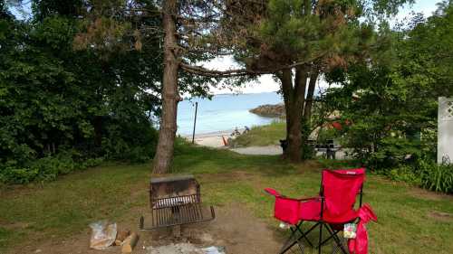 A red camping chair beside a fire pit, with a view of a calm beach and trees in the background.