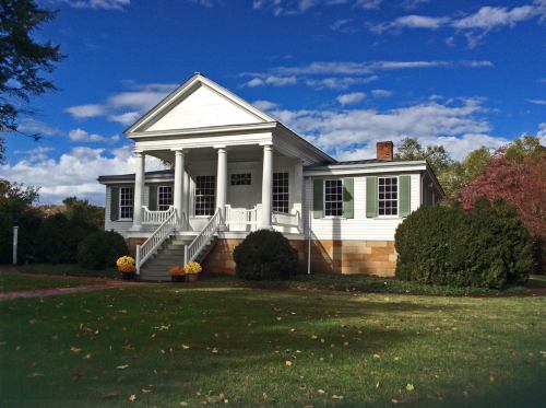A white historic house with columns, surrounded by green grass and colorful autumn foliage under a blue sky.