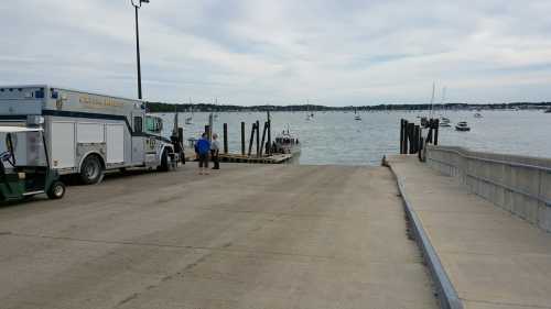 A boat ramp leads to a calm waterway with boats, alongside an emergency vehicle and two people on the dock.