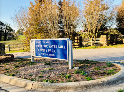 Sign for Historic Yates Mill County Park in Wake County, located on Lake Wheeler Road, surrounded by trees.