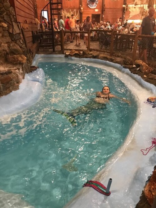 A person swims in a circular pool surrounded by stone walls, with a lively restaurant atmosphere in the background.