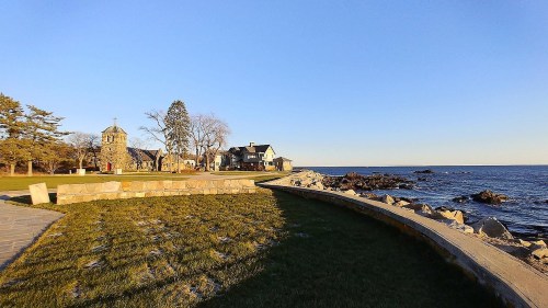 Coastal view featuring a stone building, grassy area, and rocky shoreline under a clear blue sky.