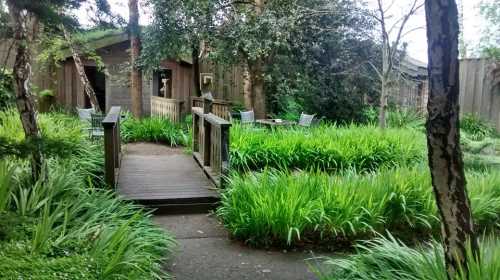 A wooden bridge leads through lush green grass to a cozy garden with chairs and a rustic building in the background.