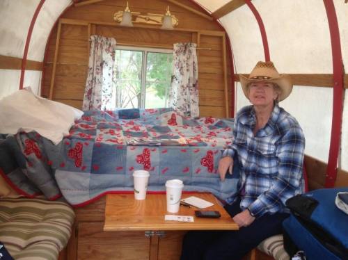 A person in a cowboy hat sits at a table inside a cozy camper, with a bed covered in a colorful quilt in the background.