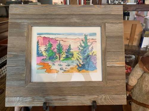 Framed watercolor painting of colorful trees and a landscape, displayed on a stand.