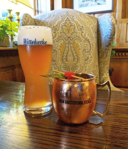 A glass of beer and a copper mug cocktail sit on a wooden table in a cozy setting.