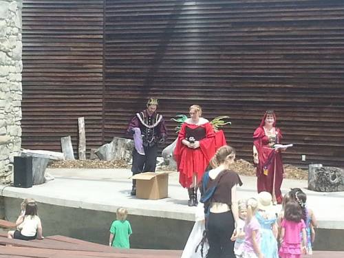 A stage performance with actors in colorful costumes, entertaining a group of children in a sunny outdoor setting.