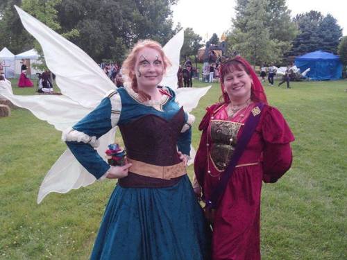Two women in colorful costumes stand in a grassy park, one dressed as a fairy and the other in a medieval gown.