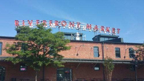 Sign for Riverfront Market atop a brick building, with trees and blue sky in the background.