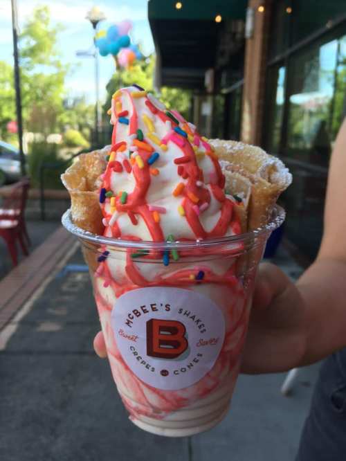 A hand holds a cup of soft serve ice cream topped with colorful sprinkles and waffle cone pieces.