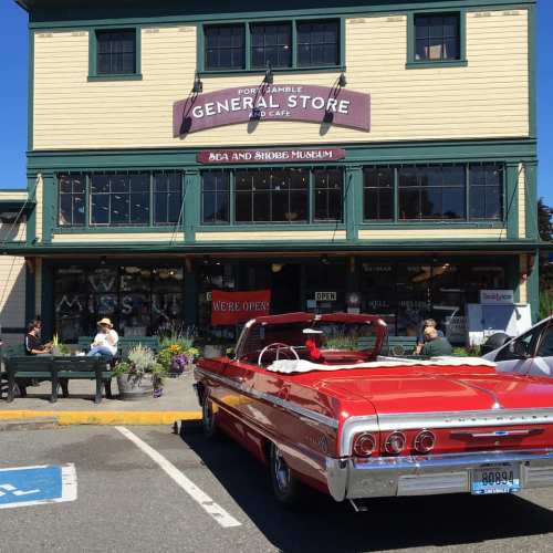 A red convertible parked in front of a general store and café with outdoor seating and a museum sign.
