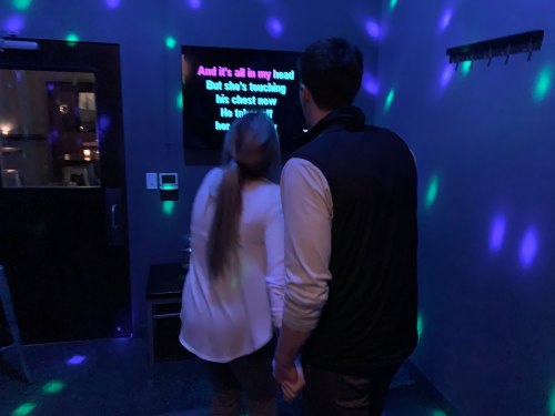 A couple stands in a dimly lit room with colorful lights, singing karaoke from a screen.