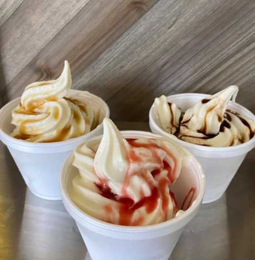 Three cups of soft serve ice cream in caramel, strawberry, and chocolate flavors, set against a wooden background.