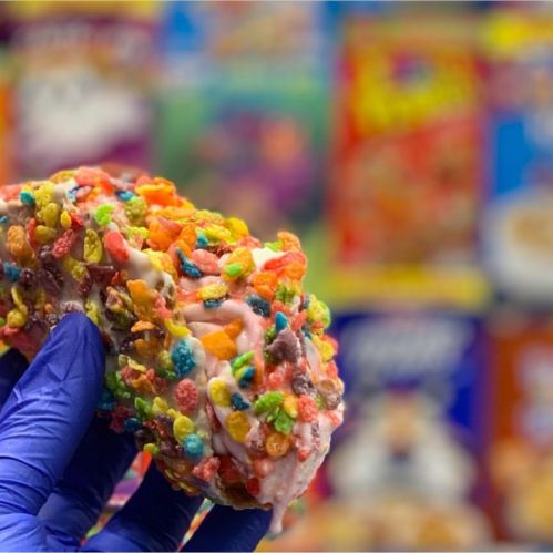 A colorful donut coated in fruity cereal, held by a gloved hand, with a vibrant cereal box background.