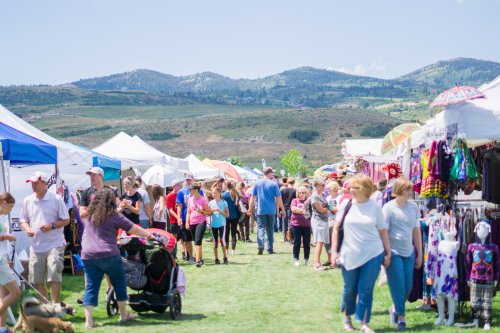 A bustling outdoor market with people browsing tents and stalls, set against a backdrop of mountains and blue skies.